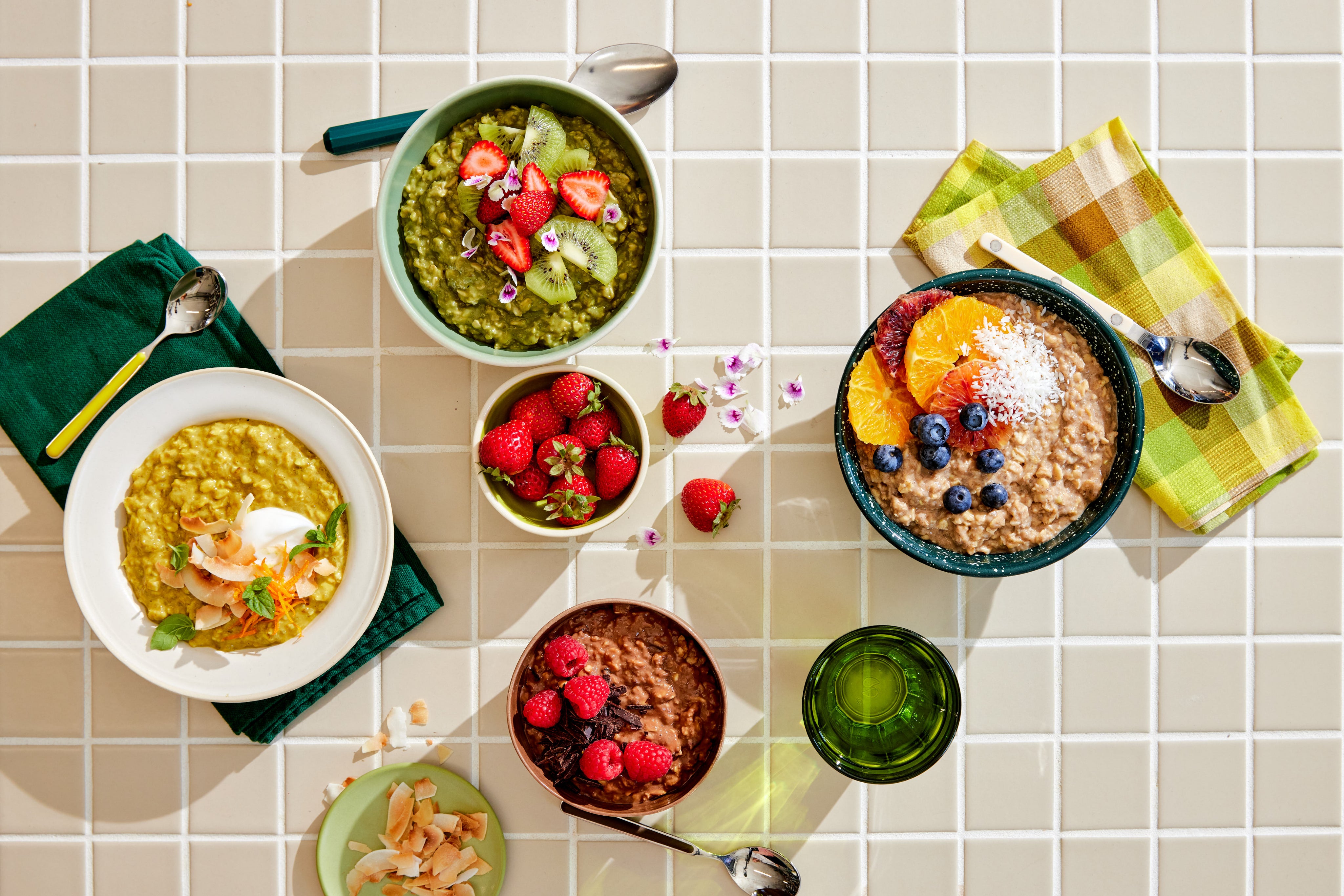 A variety of colorful breakfast bowls on a tiled surface display vibrant green and yellow porridges topped with fruits and nuts. An accompanying dish of strawberries and scattered flower petals add a decorative touch. Nearby, spoons accompany a stylish green glass, completing the vibrant scene.
