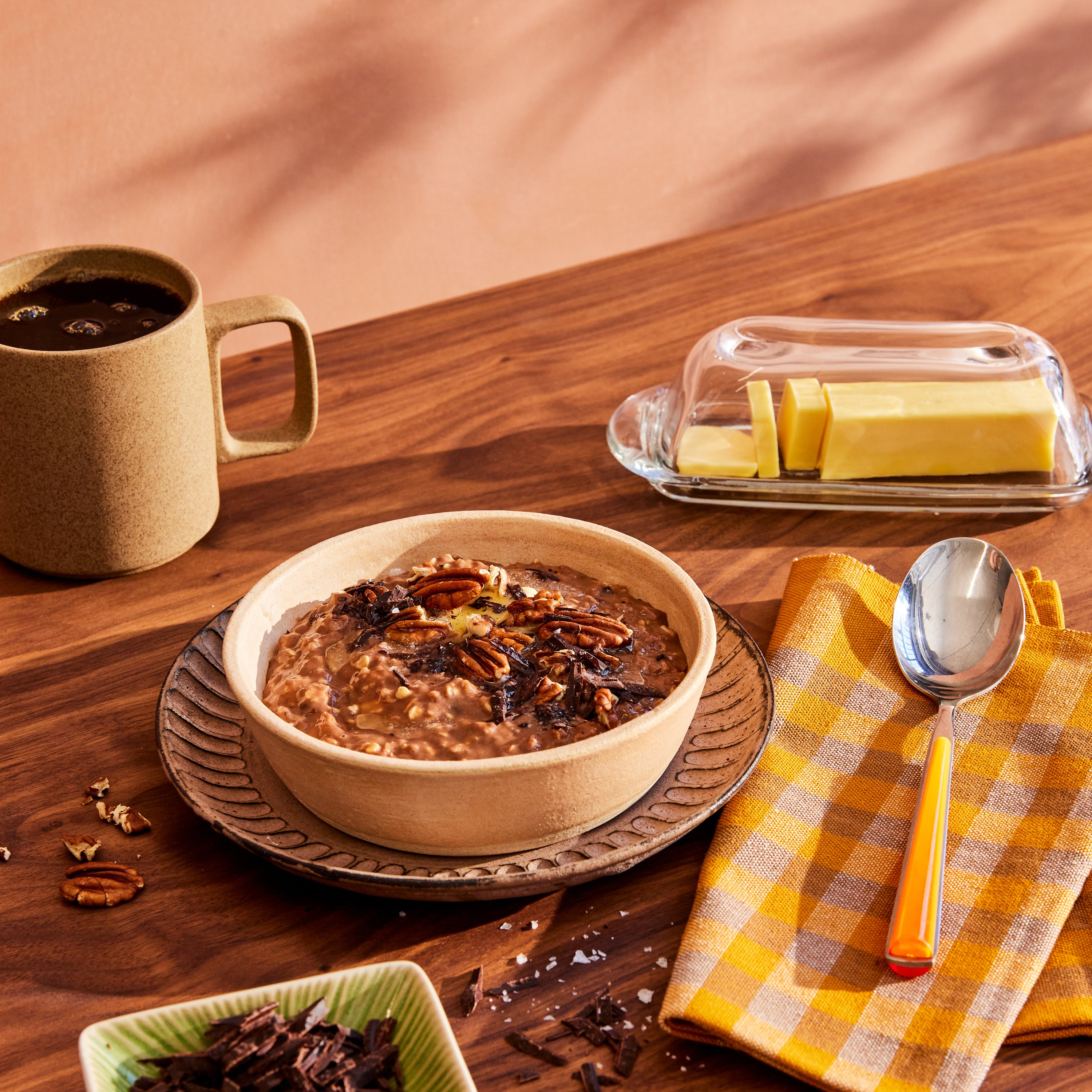 A cozy breakfast scene features a bowl of oatmeal topped with pecans and chocolate shavings on a wooden table. A spoon rests on a yellow checkered napkin next to a steaming mug of coffee and a butter dish. Gentle shadows dance across the wall behind, creating an inviting morning atmosphere.