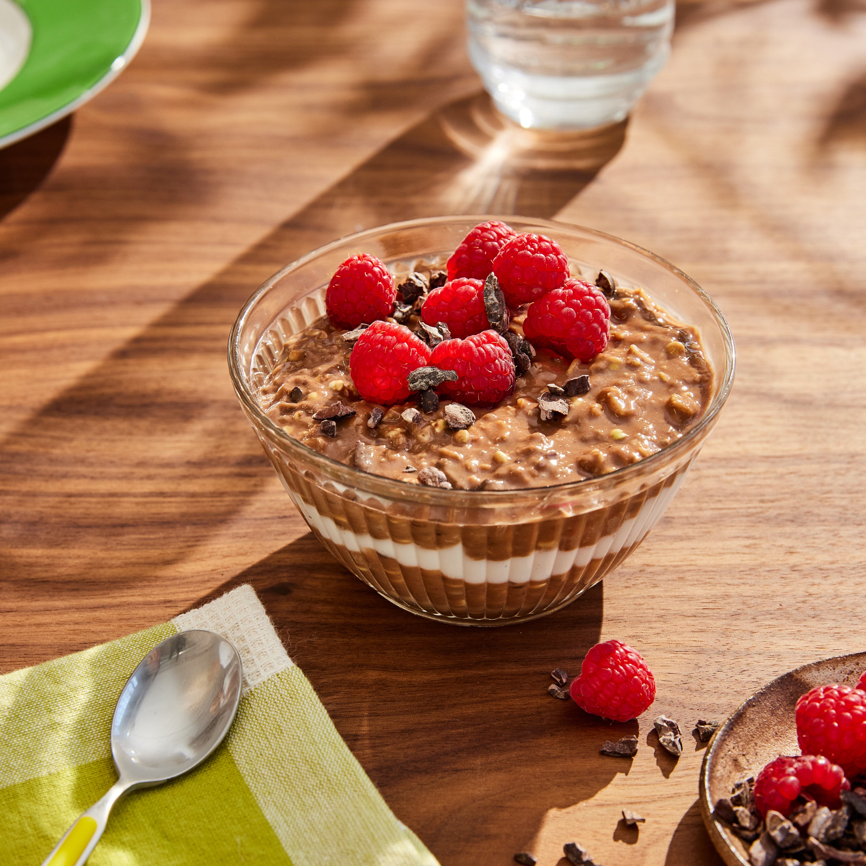 A bowl of chocolate chia pudding topped with fresh raspberries and cacao nibs sits on a wooden table. A spoon is placed next to a green and white napkin, and sunlight casts patterns across the scene.