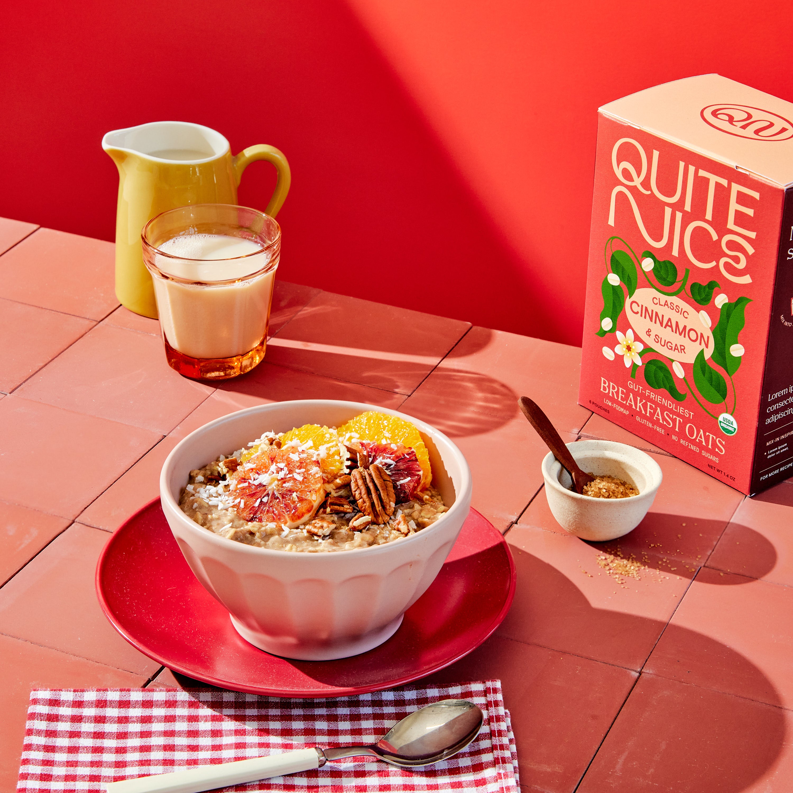 A bowl of creamy oatmeal topped with fresh orange slices, crunchy pecans, and brown sugar rests on a vibrant red plate with a checkered napkin. Nearby are a box of wholesome oats, a spoon, a small bowl of sugar, a glass of milk, and a cheerful yellow pitcher on the red tabletop.