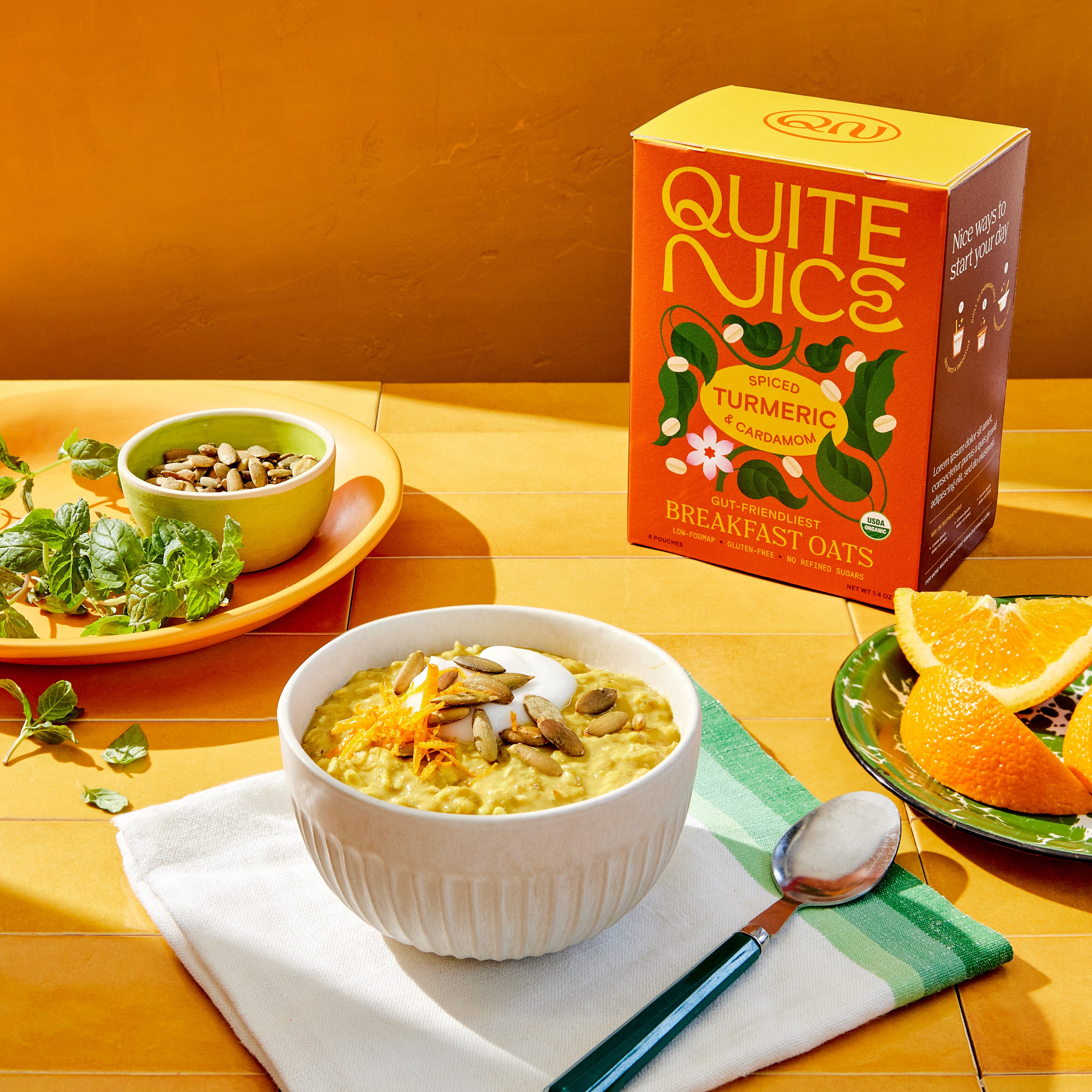 A vibrant breakfast scene features a bowl of turmeric cardamom oats topped with nuts and a spoon on a cloth. In the background, a box labeled Quite Nice oats sits alongside fresh mint, seeds in a bowl, and sliced oranges on a plate.