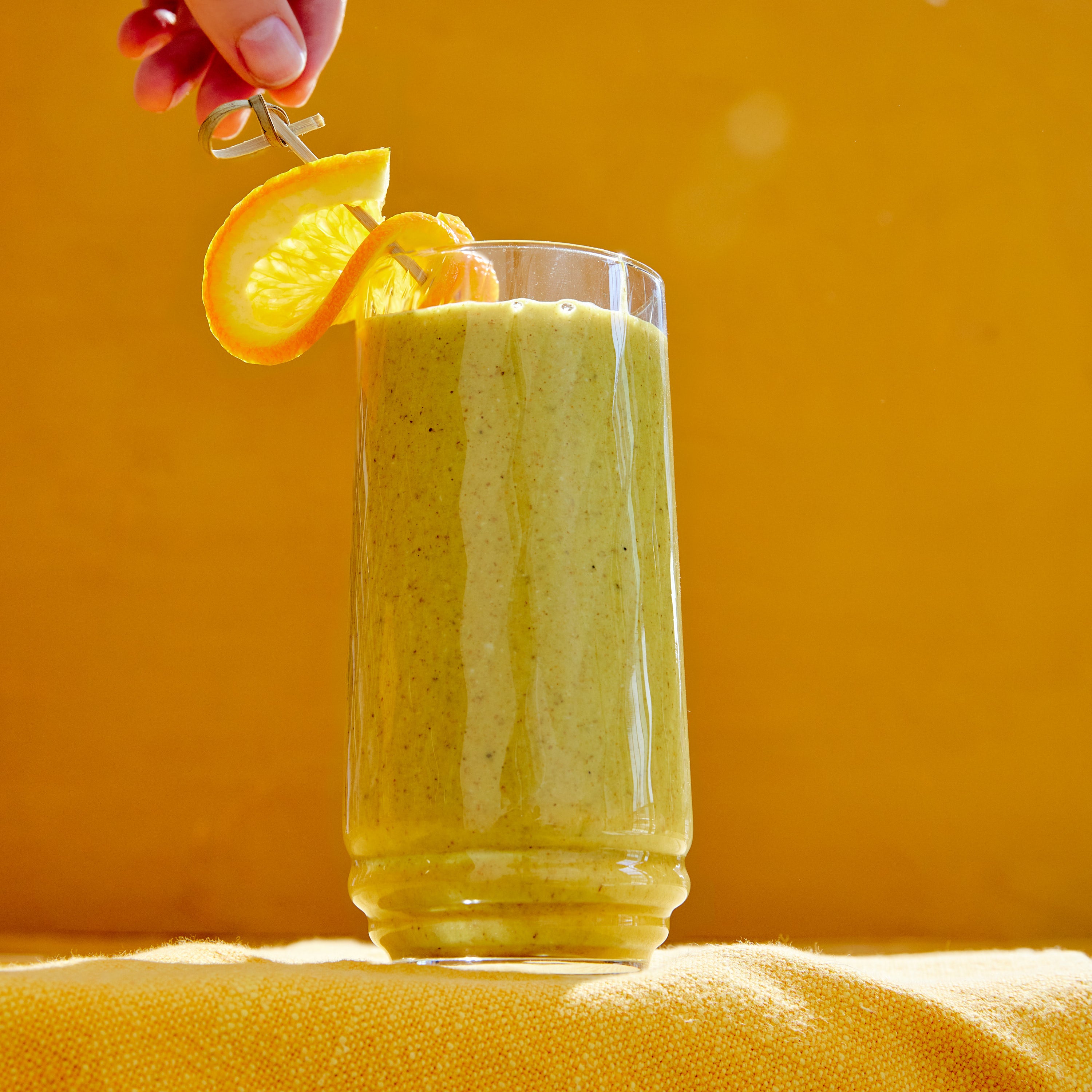 A tall glass of green smoothie garnished with an orange slice on a skewer against a bright yellow background. A hand is holding the skewer, casting a soft shadow on the smoothies surface.