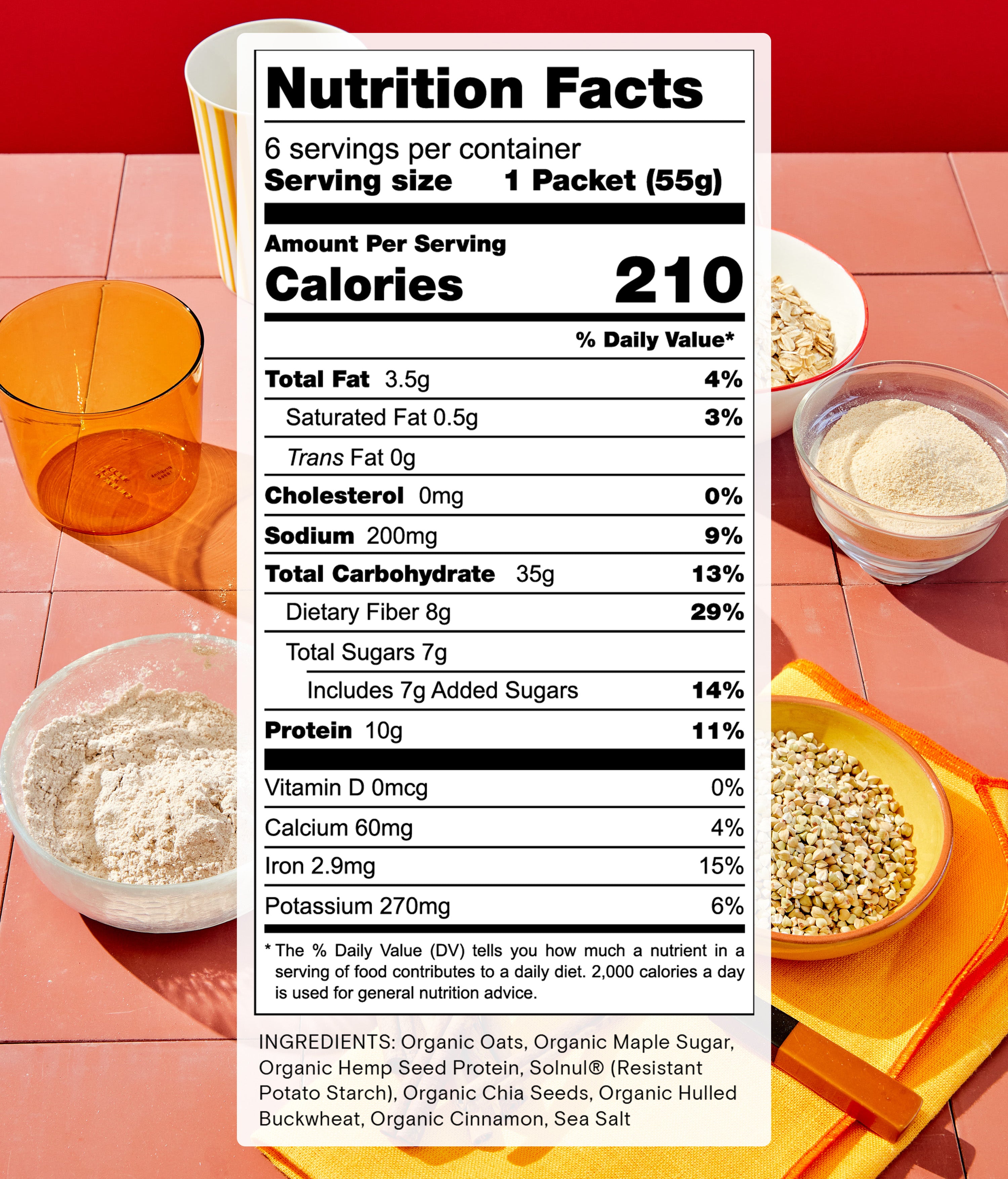 A box of oatmeal sits on a coral-colored table next to a white mug and a glass filled with orange juice. Other bowls contain oats and a brown sugar mix. The nutrition facts and ingredients are displayed prominently in a label on the image.