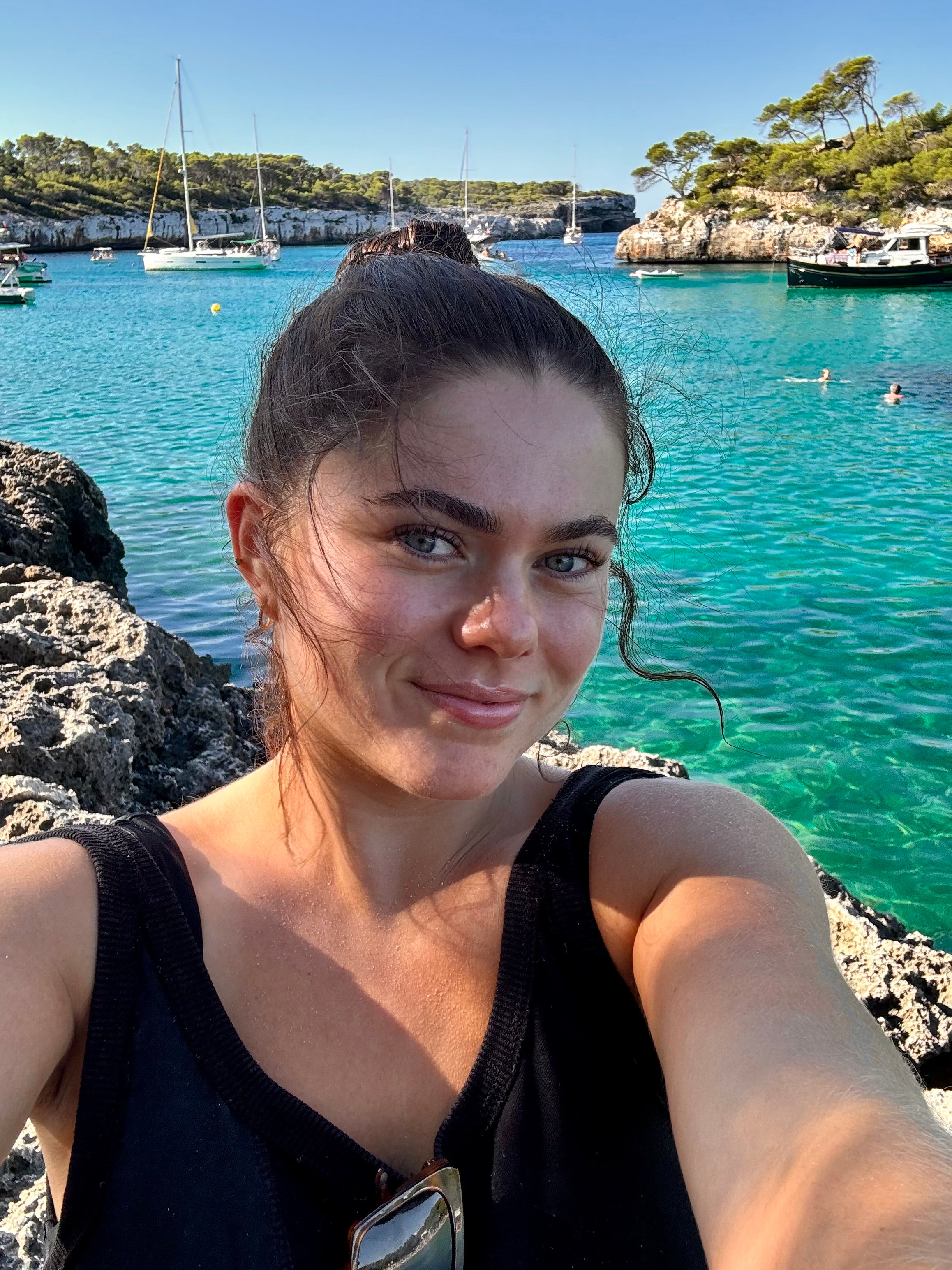 A person taking a selfie in front of a turquoise bay with rocky cliffs and sailboats. Clear blue sky and people swimming in the background.