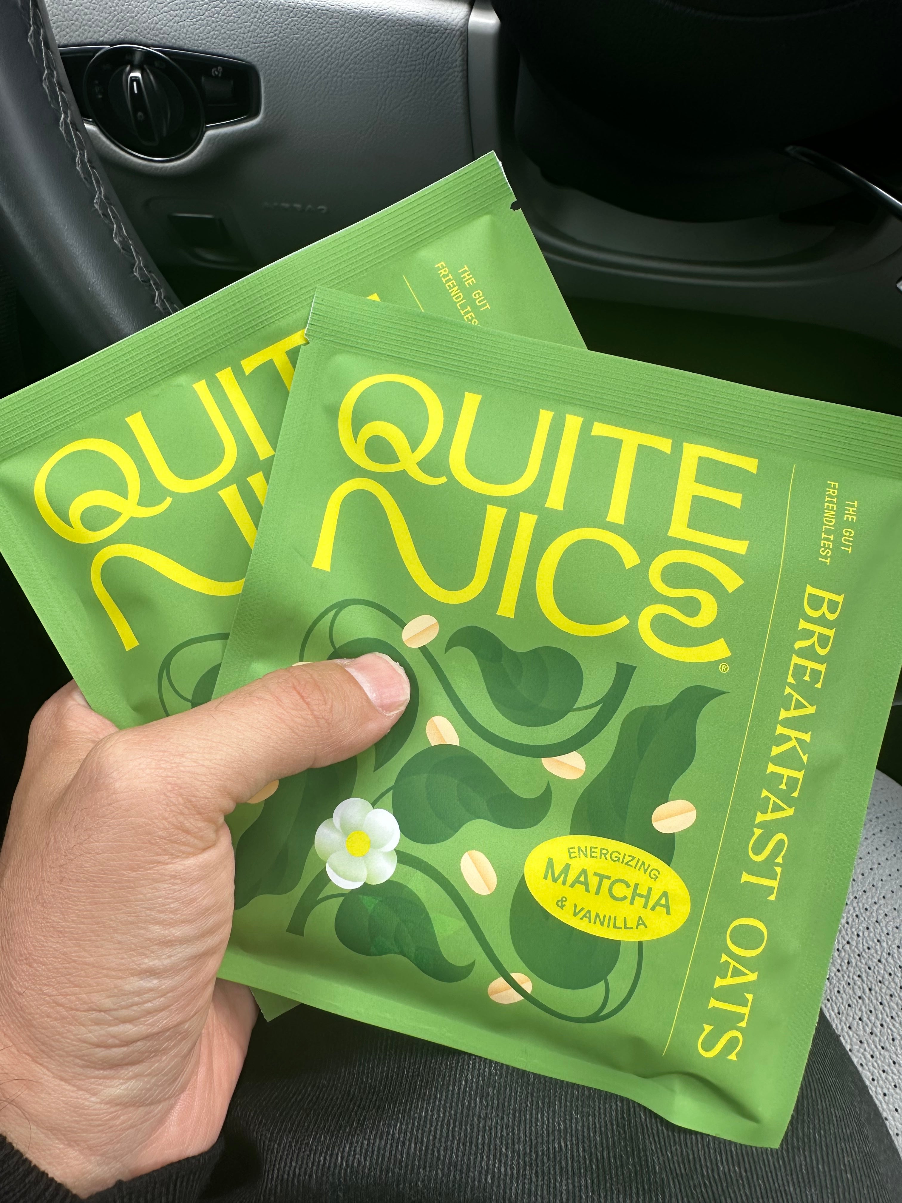 A hand holding two green packets of Quite Nice breakfast oats, labeled with Energizing Matcha & Vanilla and featuring a decorative design with leaves and a flower. The background shows part of a car interior.