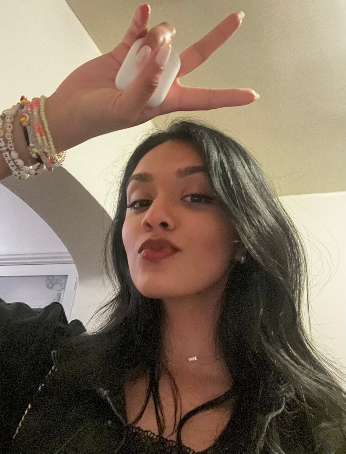 A woman with long dark hair is making a peace sign with her hand raised. She is wearing a black jacket, layered necklaces, and colorful bracelets. She looks at the camera with a slight smile against a neutral indoor background.