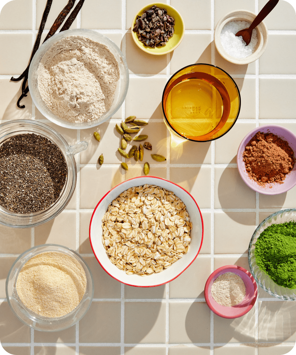 An assortment of baking ingredients on a tiled surface, including oats, flour, cocoa powder, cardamom pods, salt, chia seeds, matcha powder, chocolate chips, cornmeal, and a bowl of golden liquid.