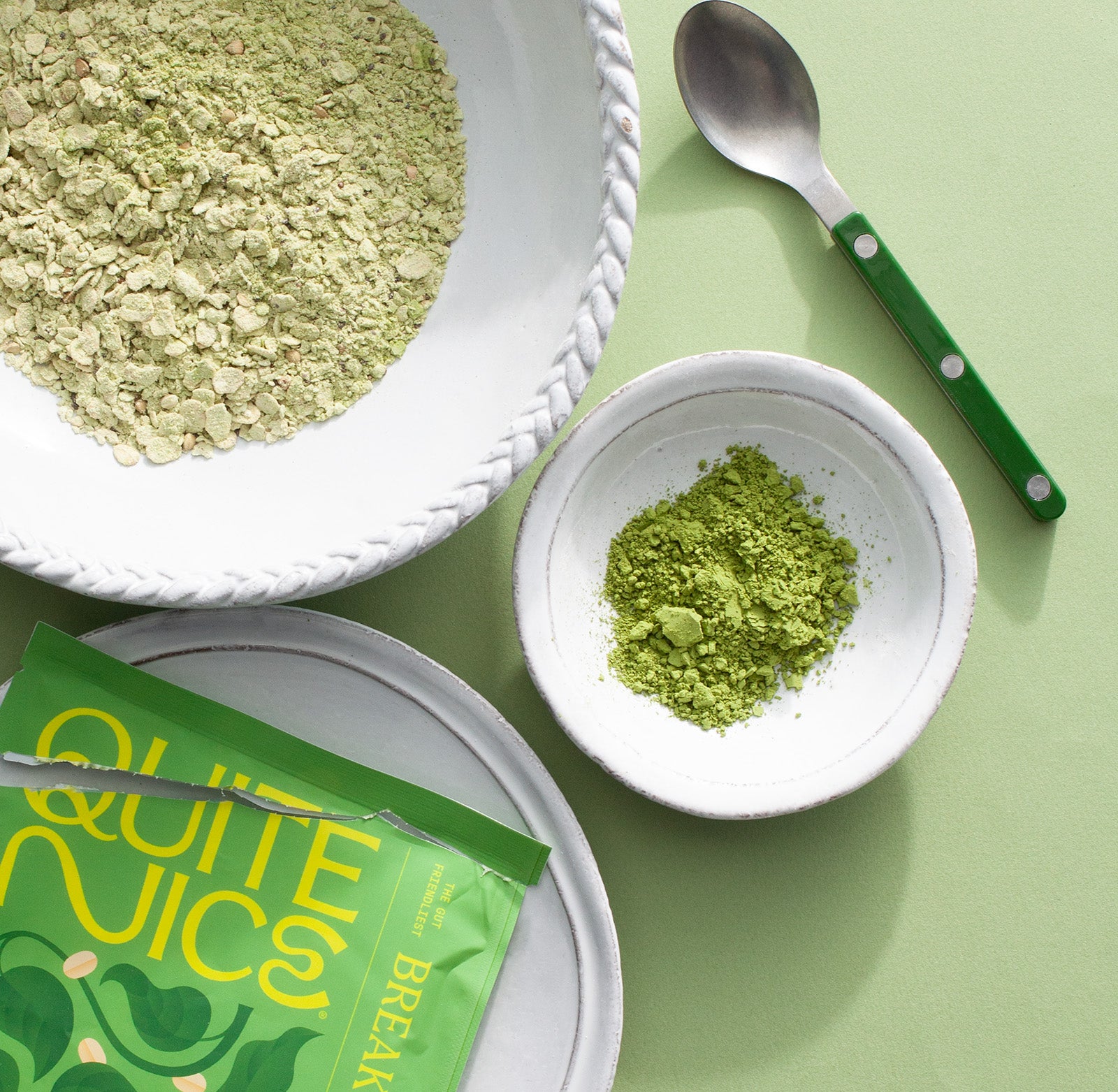 A large bowl of green cereal sits on a light green surface next to a smaller bowl of green powder. A silver spoon with a green handle and a green Quite Nice cereal package are also visible.