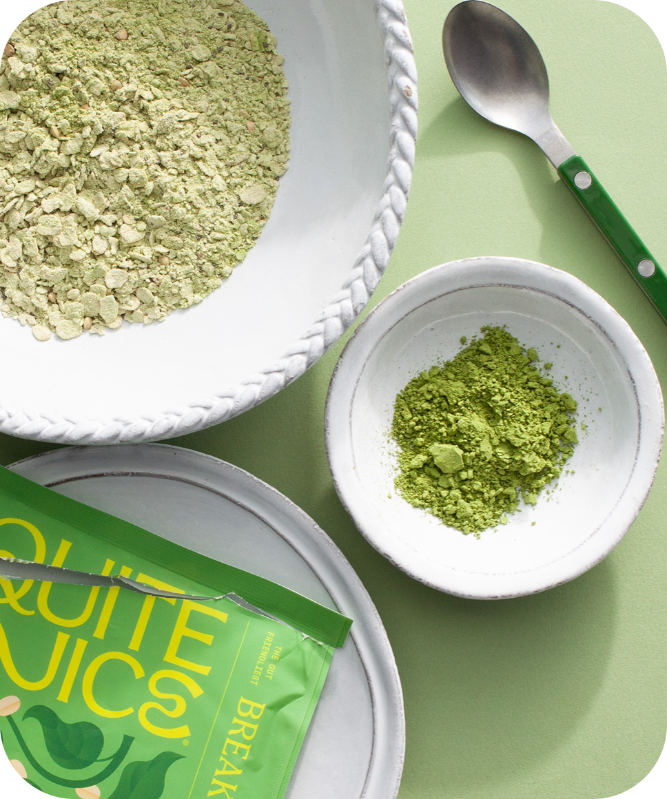 A green smoothie powder in a package, alongside a bowl filled with green flakes, a smaller bowl with finely ground green powder, and a spoon with a green handle. All items are placed on a light green surface.