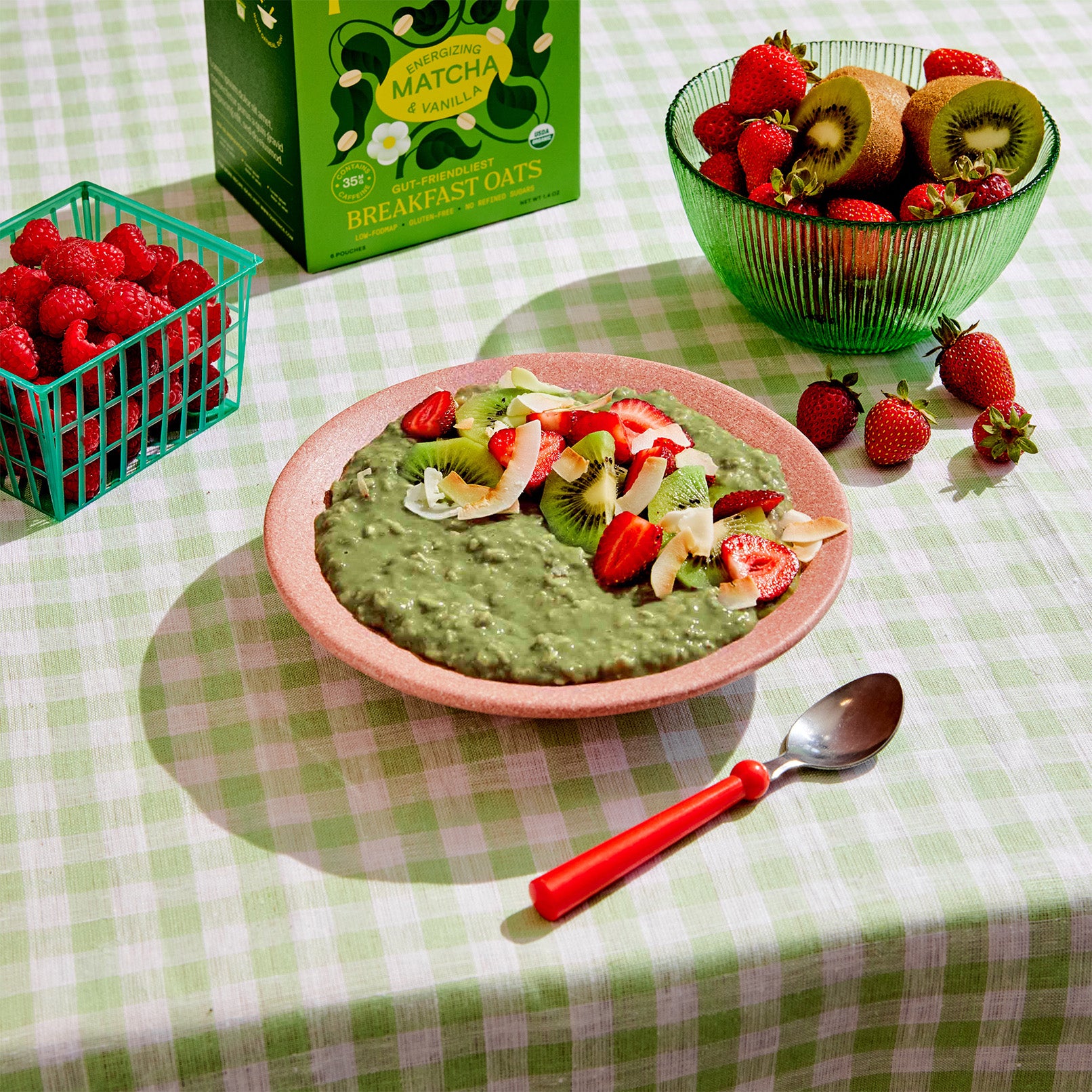 A vibrant bowl of green matcha oatmeal adorned with sliced strawberries, kiwi, and coconut flakes sits on a checkered tablecloth. A spoon rests beside it, accompanied by containers of fresh raspberries, strawberries, and kiwis. A box of aromatic matcha oats stands nearby.