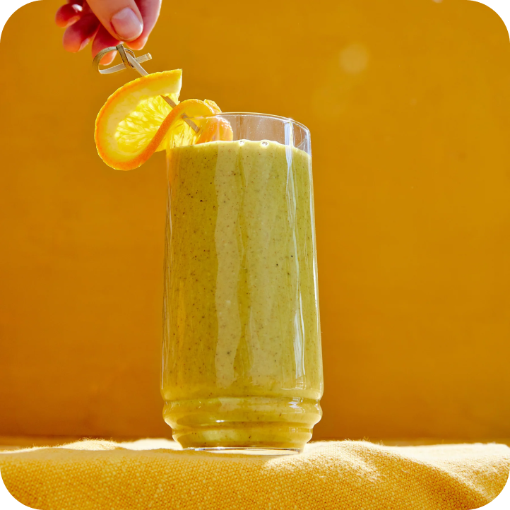 A tall glass of creamy green smoothie, crafted with real food ingredients for optimal gut health, is garnished with an orange slice and a metal skewer against a vibrant yellow background. A hand elegantly holds the skewer, adding a decorative touch.