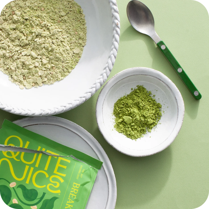 Top view of a green-themed setup featuring a packet labeled QUITE NICE, a bowl of gut health-boosting matcha powder, a larger bowl with fiber-rich granola, and a green-handled spoon, all placed on a light green background.