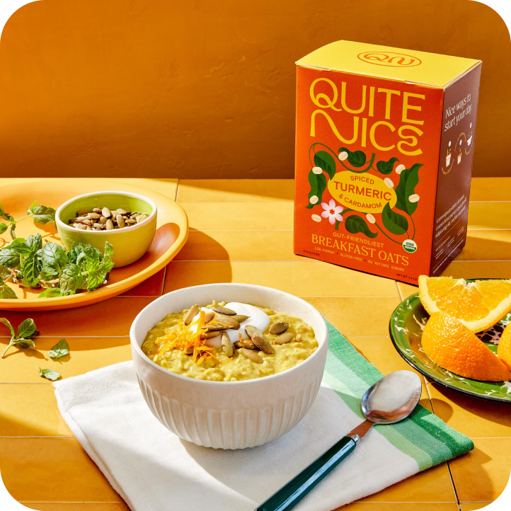 A bowl of spiced turmeric cardamom oats, made with real food ingredients, is topped with nuts on a white napkin with a spoon. Nearby, a small dish holds seeds, and a plate has orange slices. A mint garnish and a box of Quite Nice oats complete the setup on an orange surface.