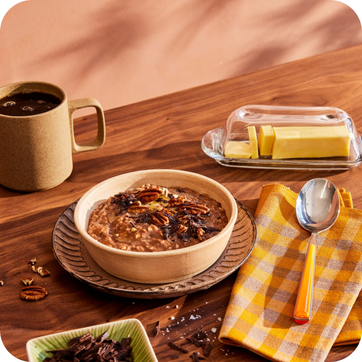 A bowl of oatmeal, rich in fiber, topped with pecans and chocolate on a wooden table, accompanied by a cup of coffee and a butter dish with butter slices. A spoon rests on a yellow checkered napkin beside the bowl—a perfect start for your gut health routine.