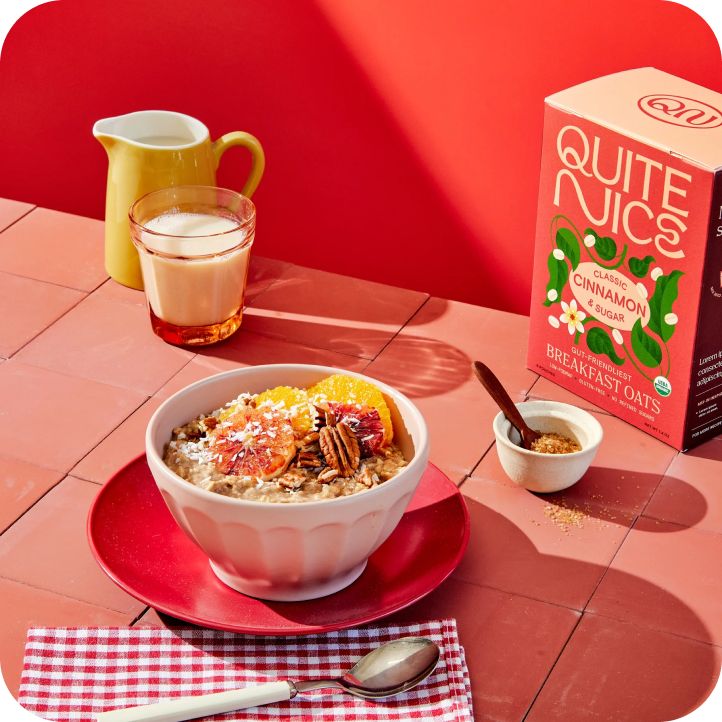 A bowl of oatmeal topped with oranges, nuts, and fruit rests on a red plate with a spoon on a checkered napkin. Nearby are a cup of milk, a small jug, brown sugar, and Quite Nice Breakfast Oats—perfect for adding fiber to your routine. The background is a red tiled surface.