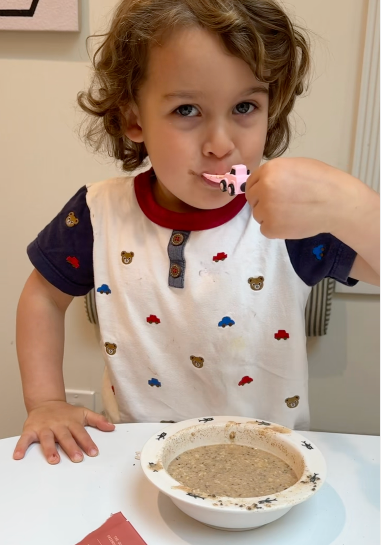 A young child with curly hair enjoys a spoonful of oatmeal, standing at a table. They're wearing a shirt adorned with bear and cloud patterns, adding a playful touch to their morning routine.