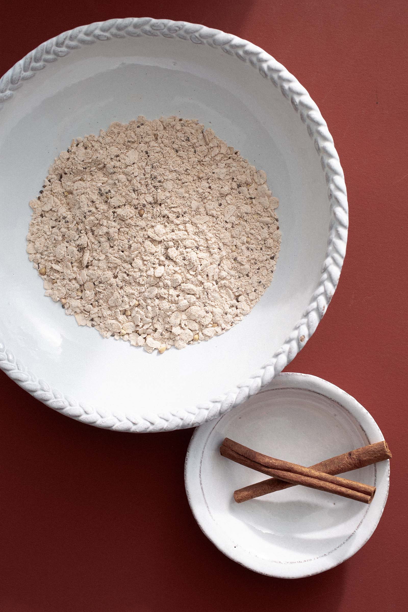 A white bowl filled with oats is placed on a red surface. Next to it, a smaller white dish holds two cinnamon sticks. The dishes are simple with decorative edges, creating a minimalistic and natural aesthetic.