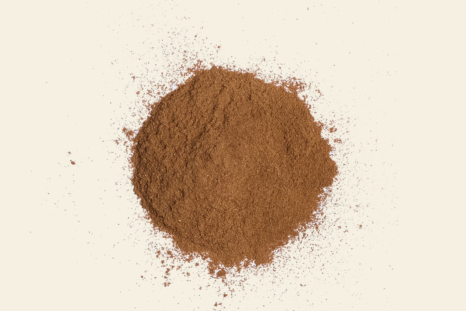 A mound of finely ground cinnamon powder is spread on a plain light-colored background. Sprinkled with earthy warmth, the aromatic spice forms a round shape with some loose particles artistically scattered around the edges.