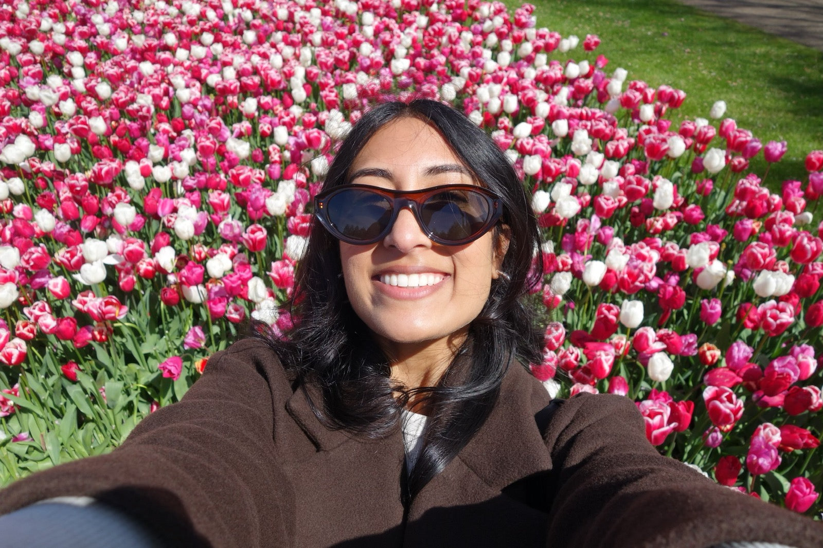 A person wearing sunglasses and a brown coat takes a selfie in front of a vibrant field of pink and white tulips under a sunny sky.