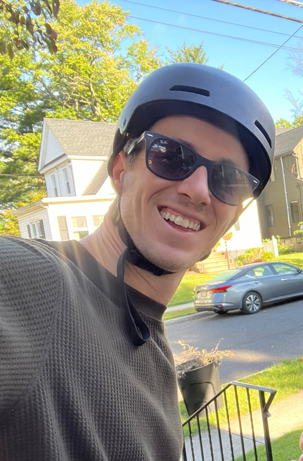 A person wearing a gray helmet and sunglasses smiles, standing outside on a sunny day. Theres a house and a parked car in the background, with trees and a clear blue sky.