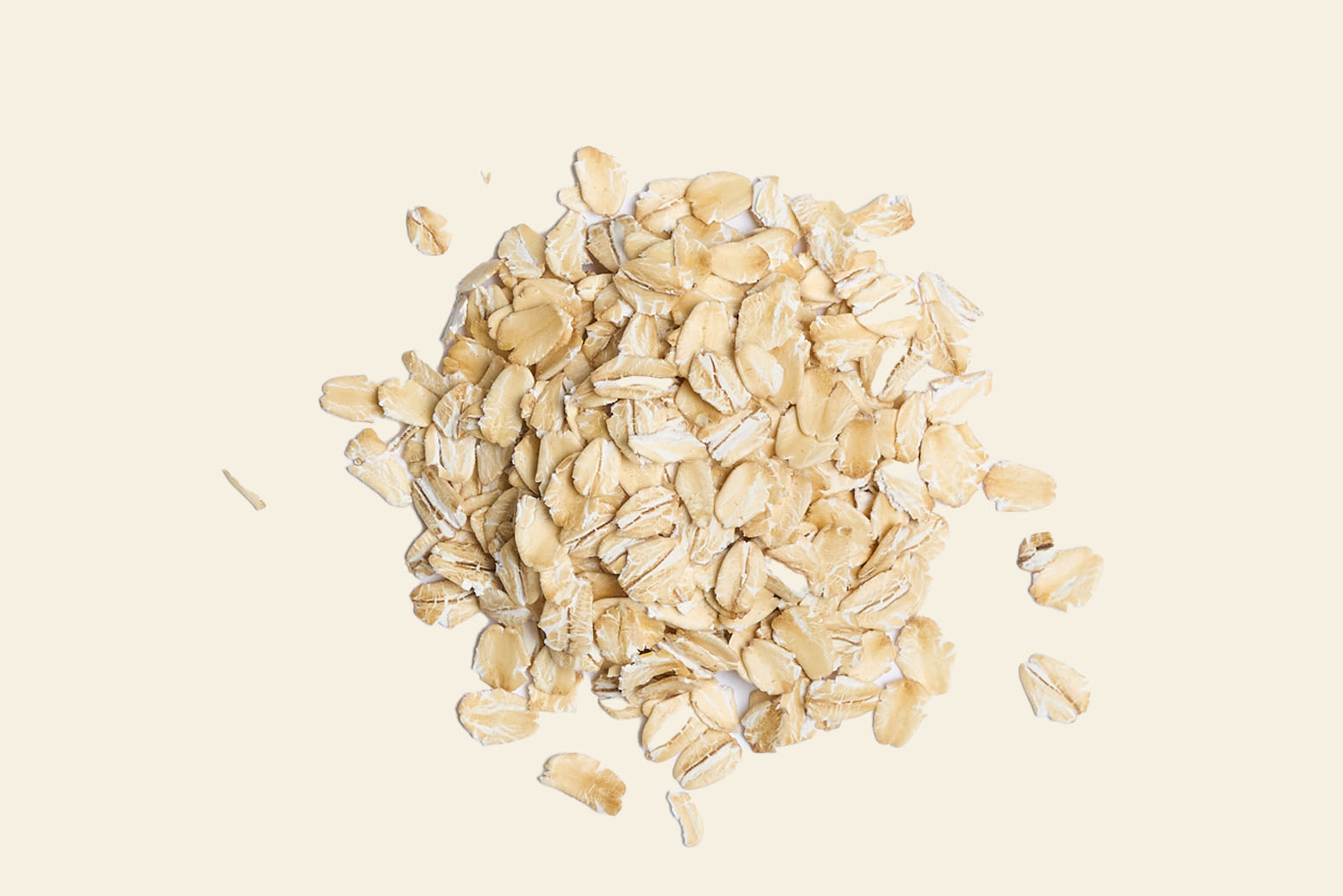 A pile of uncooked rolled oats rests on a plain beige background, with the whole grains slightly scattered around the edges.